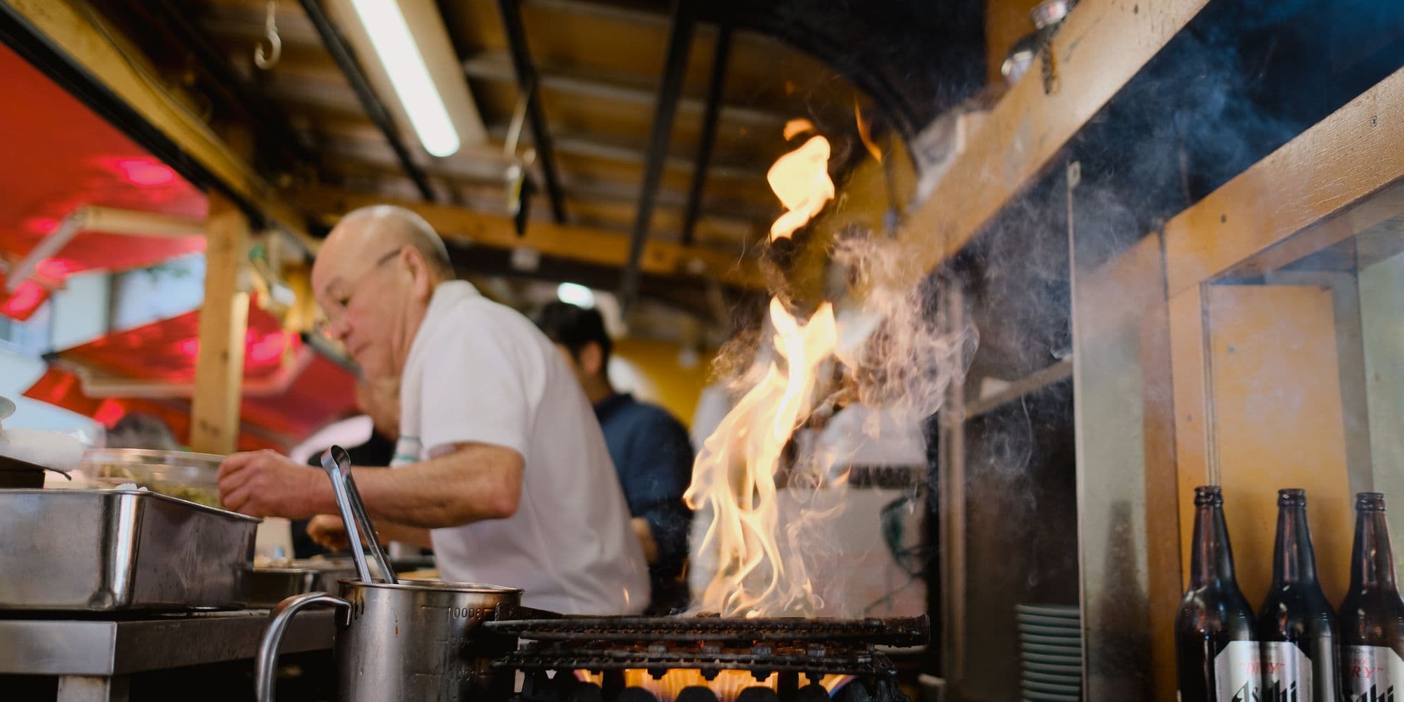 The open kitchen at Ember & Bun Syntagma during dinner service, flames rising from the grill